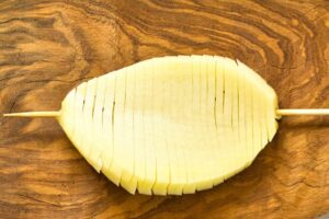 A skewer inserted into an accordion potato laying on a cutting board.