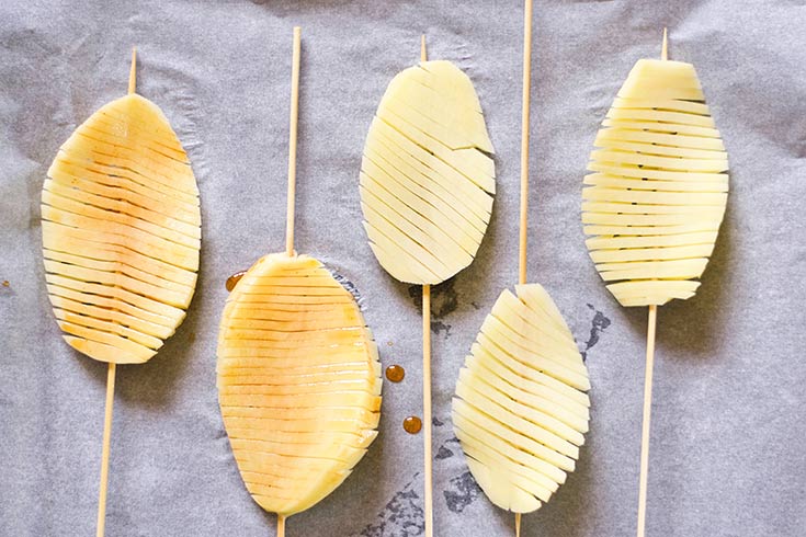 Dried off accordion potatoes on skewers, based with oil and laying on a parchment-lined cookie sheet.