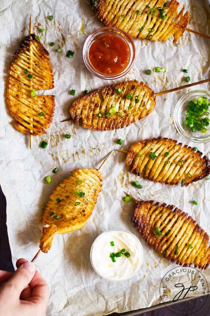 A female hand reaches for an accordion potato skewer that lays on a parchment-linked cookie sheet.