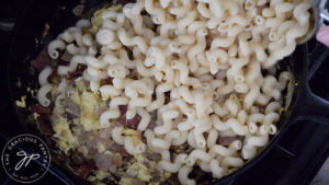 Bacon and pasta being added to sautéd onions and garlic in a cast iron skillet.