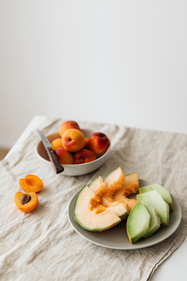 A plate of sliced melon lays on an off white tablecloth. A bowl of apricots sits behind and to the left of it.
