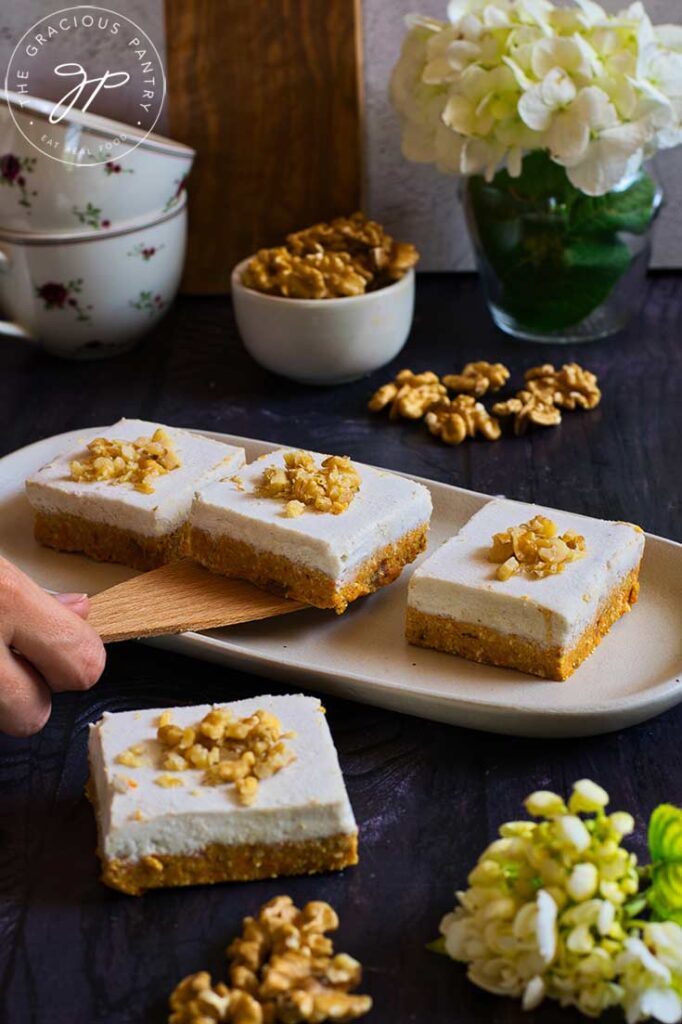 A female hand uses a wooden spatula to pick up a piece of this Gluten-Free Carrot Cake from a serving platter.