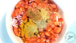 Spices added to a white mixing bowl that holds chopped veggies and chickpeas.