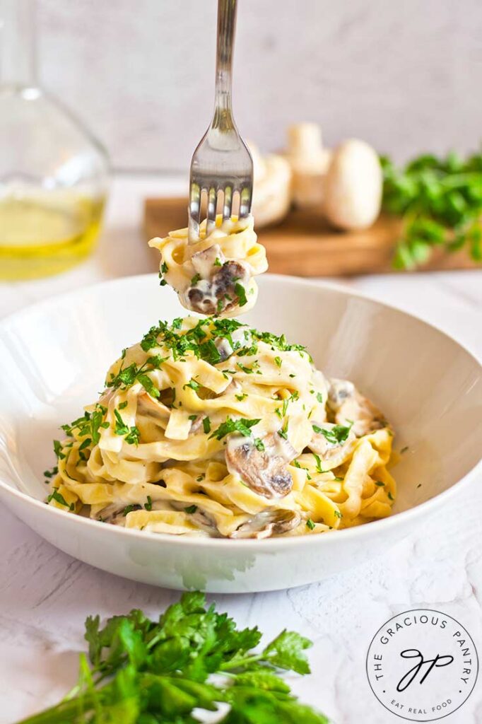 A fork holds a twirl of this vegan fettuccine alfredo pasta over a white bowl that holds a serving of the same.