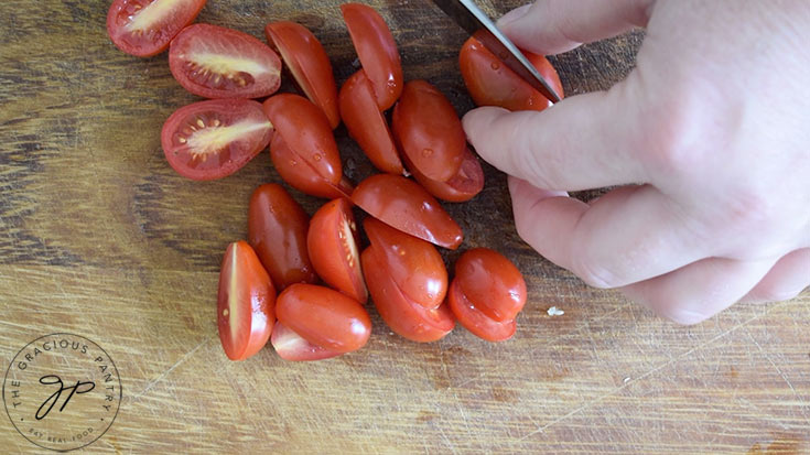 Grape tomatoes being cut in half on a wood cutting board.