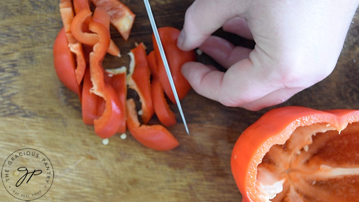 A red bell pepper being sliced on a wood cutting board.