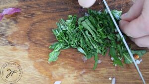 Fresh basil leaves being sliced thin on a wood cutting board.