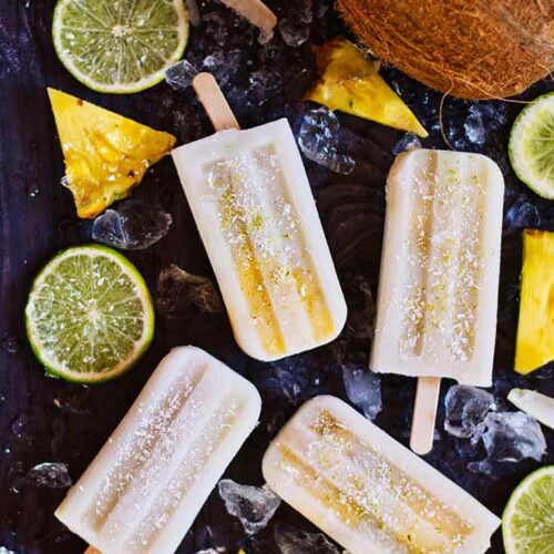 Pineapple Popsicles lay on a dark surface surrounded by lime slices and pineapple wedges.