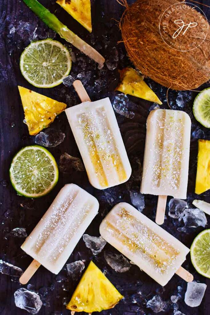 Pineapple Popsicles lay on a dark surface surrounded by lime slices and pineapple wedges.