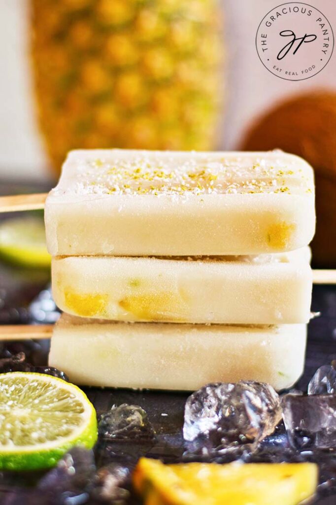 A stack of three Pineapple Popsicles is stacked up on a dark gray surface. Ice cubes surround it.