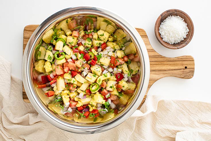 The pineapple salsa, mixed in a mixing bowl, and sitting on a cutting board against a white surface.