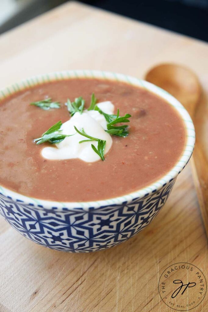 Side view of a decorative bowl filled with Refried Bean Soup, garnished with plain yogurt and fresh cilantro. A spoon rests to the right of the bowl.