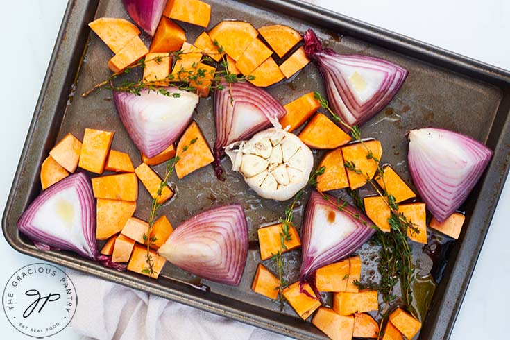 The onions, garlic and sweet potatoes with herbs and oil, sitting on a baking sheet.