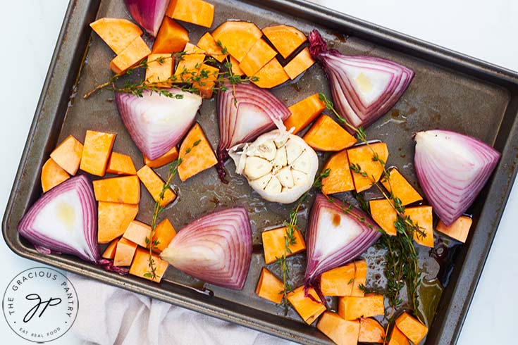 The onions, garlic and sweet potatoes with herbs and oil, sitting on a baking sheet.