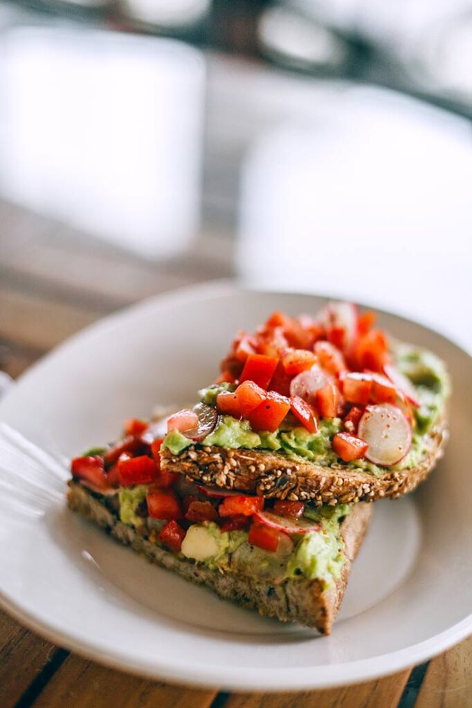 A white plate holds and open-faced sandwich with avocado and chopped tomatoes on it.