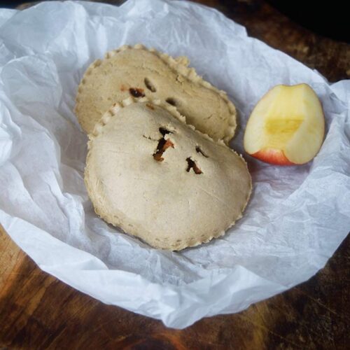 Two hand pies and a quarter of an apple lay on the piece of parchment paper on a wooden surface..