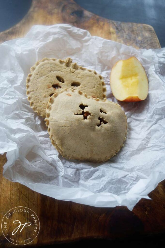 Two baked apple hand pies and a piece of apple laying on some parchment.