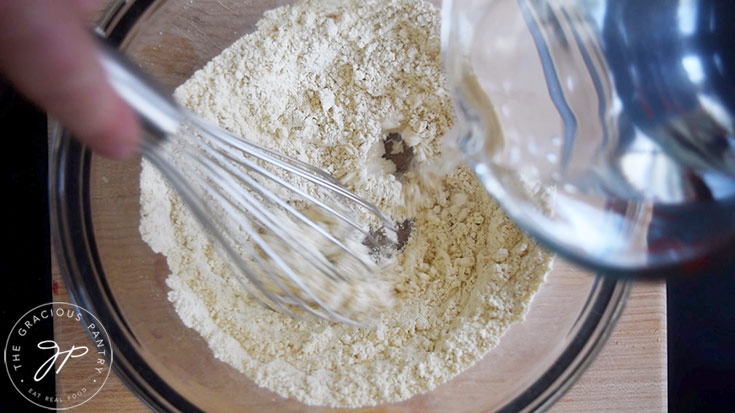 Pouring water into a glass mixing bowl that has chickpea flour and salt in it.