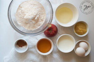 All the ingredients for Cinnamon Apple Muffins measured and sitting in individual bowls on a white surface.