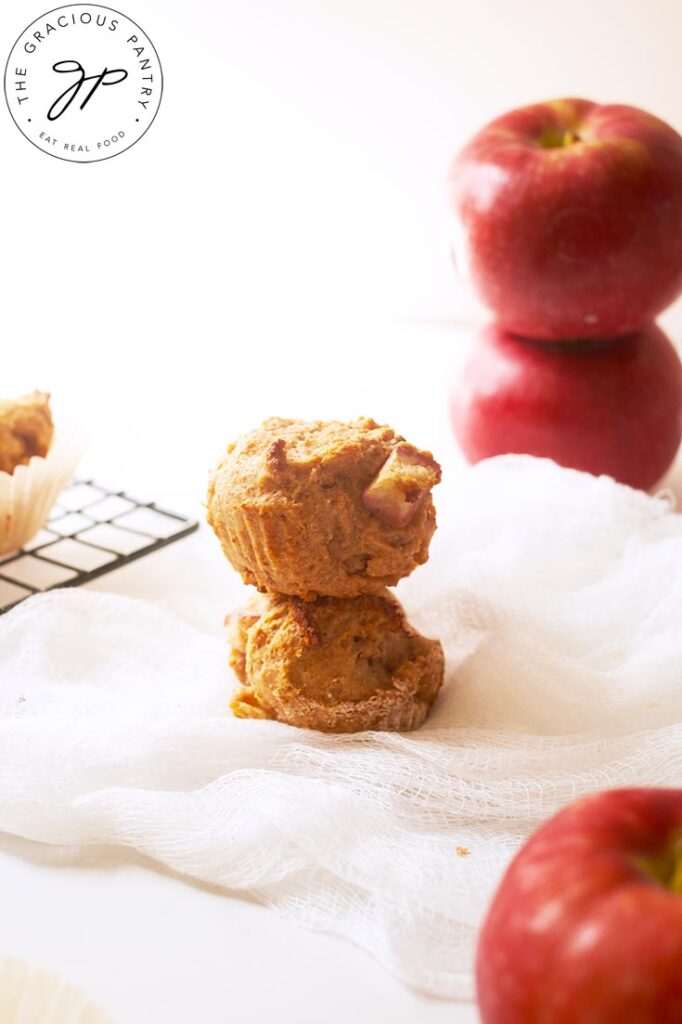 Two cinnamon apple muffins stacked up on a white surface next to some apples.