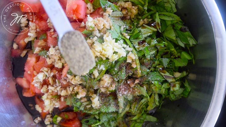 Adding spices to a mixing bowl holding chopped tomatoes, basil and garlic.