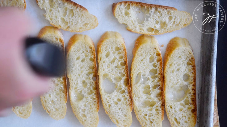 Bread slices on a cookie sheet being sprayed with an oil sprayer.