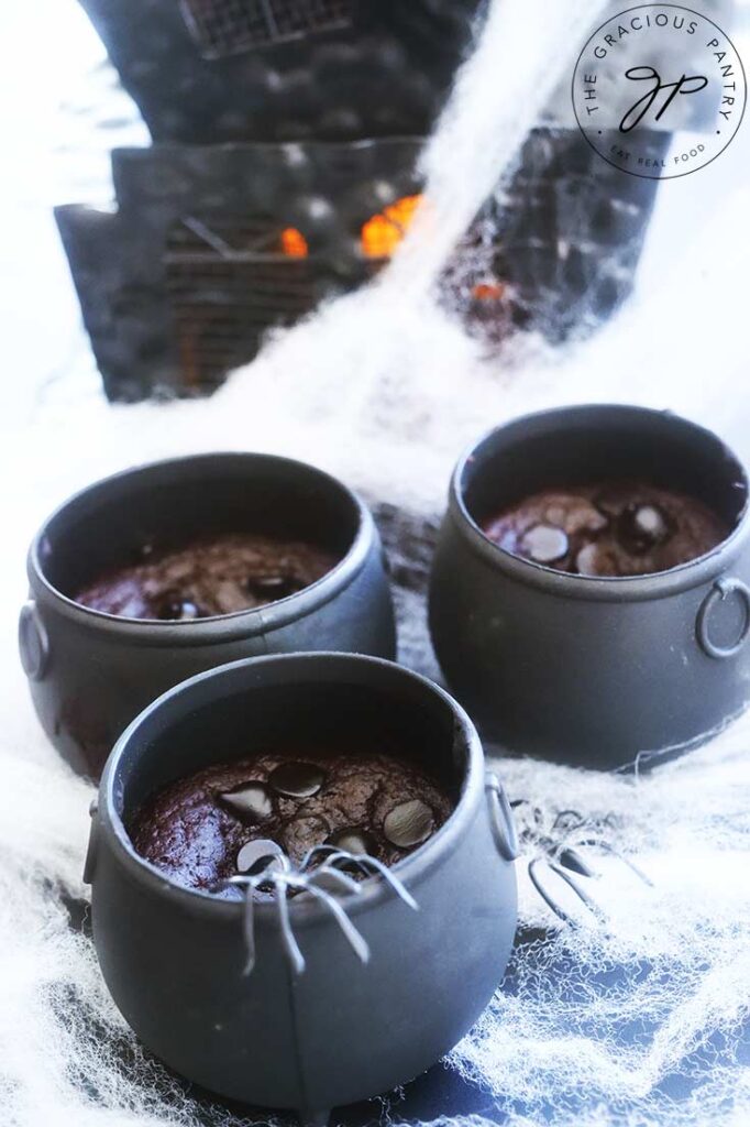 Three cauldron cakes sit in front of a black haunted house. A black spider rests on one of the cauldrons.