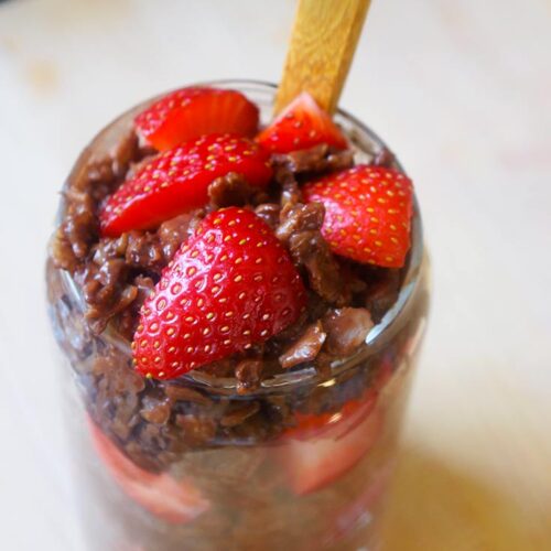 A spoon resting in a full cup of chocolate oatmeal topped with fresh cut strawberries.