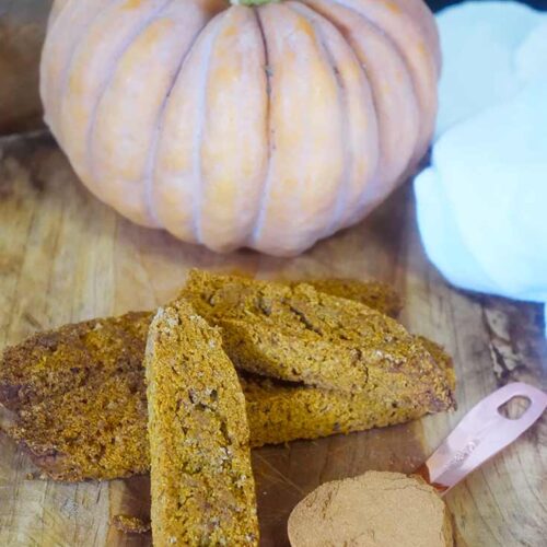 Gluten-Free Pumpkin Biscotti on a cutting board with a pumpkin and a tablespoon of pumpkin pie spice to the side.