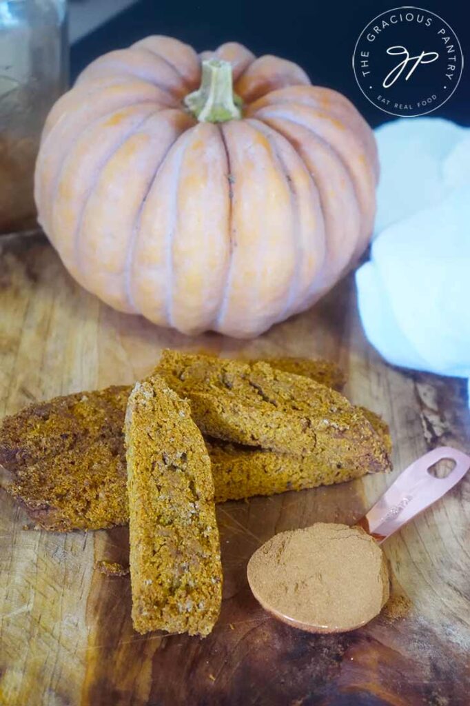 Gluten-Free Pumpkin Biscotti on a cutting board with a pumpkin and a tablespoon of pumpkin pie spice to the side.