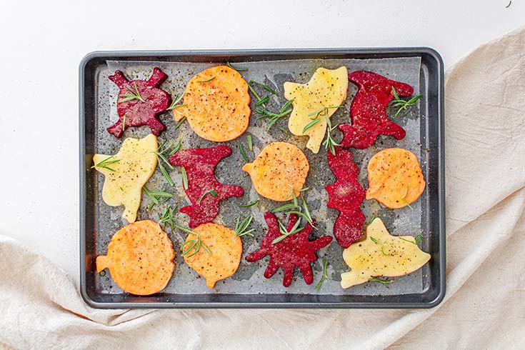 Halloween Roasted Vegetables laying on a parchment-lined baking sheet, topped with seasonings and fresh herbs.