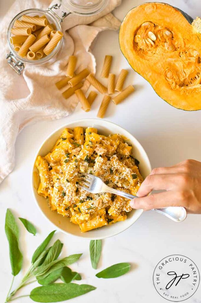 A female hand holding a fork reaches for a bite of pumpkin pasta from a white bowl.