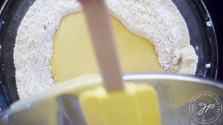 Wet egg mixture being poured into dry flour mixture in a glass mixing bowl.