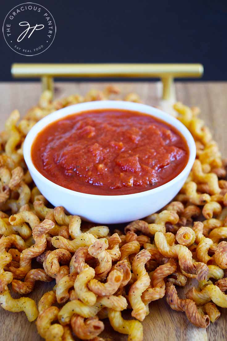 A white bowl of marinara sits on a wooden board surrounded by Air Fryer Pasta Chips.