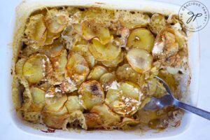 A spoon taking a serving of Healthy Scalloped Potatoes our of a white casserole dish.