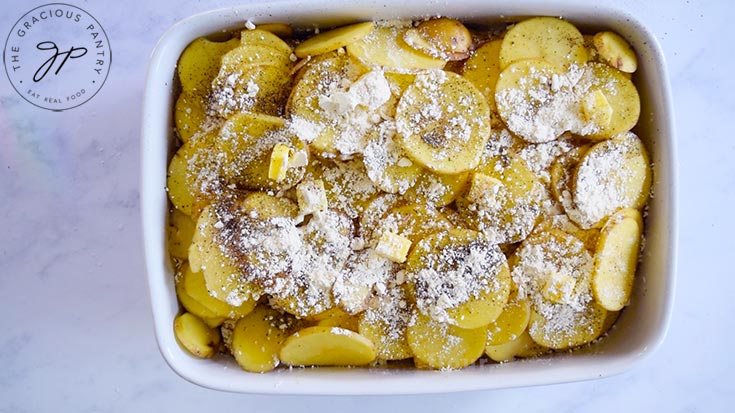 Sliced potatoes in a white casserole dish topped with spices, flour and butter.