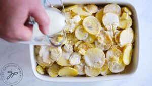 Pouring milk over sliced potatoes in a white casserole dish.