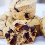A stack of Pancake Muffins sit behind a single Pancake Muffin that has been cut in half showing all the chocolate chips inside.