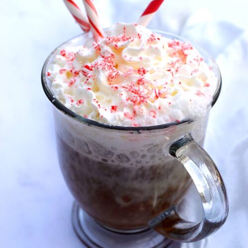 A glass mug filled with peppermint latte and topped with whipped cream, crushed candy cane and a whole candy cane for garnish.