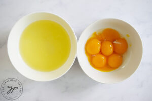 Two white bowls on a white surface. Once has egg whites, the other egg yolks.