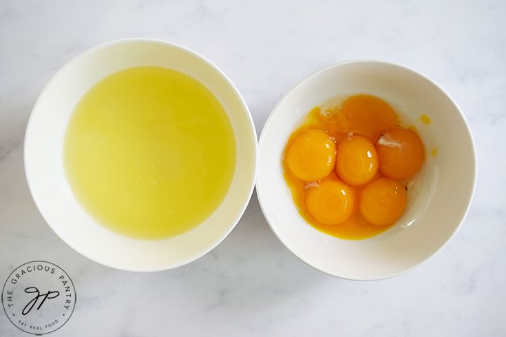 Two white bowls on a white surface. Once has egg whites, the other egg yolks.