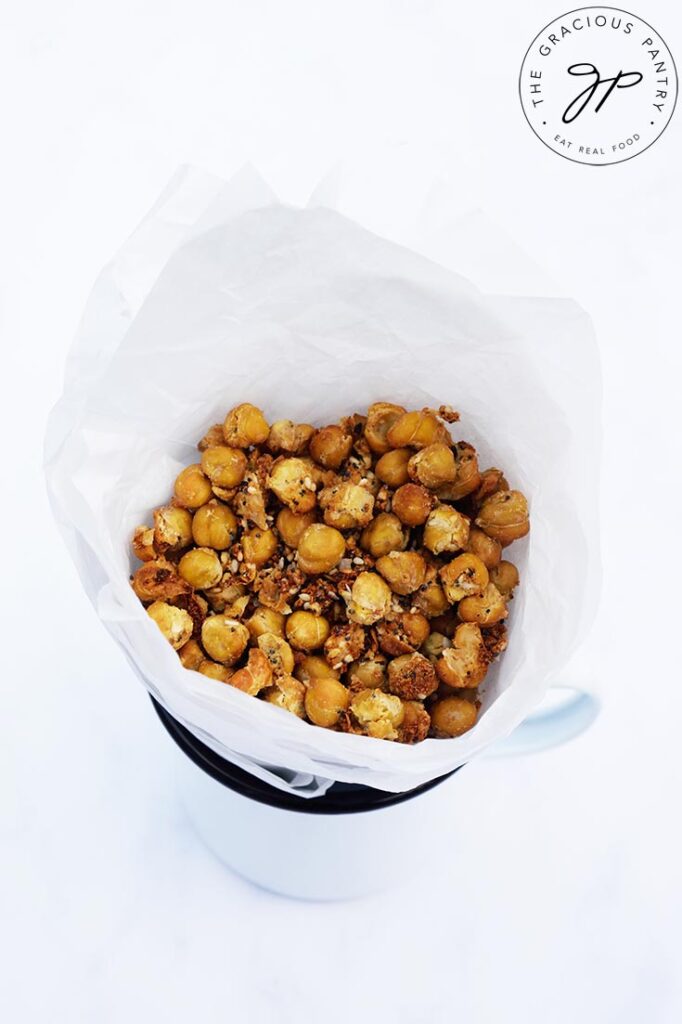 A side view of a parchment cone filled with Roasted Everything Bagel Chickpeas, sitting in a white mug with a black rim.