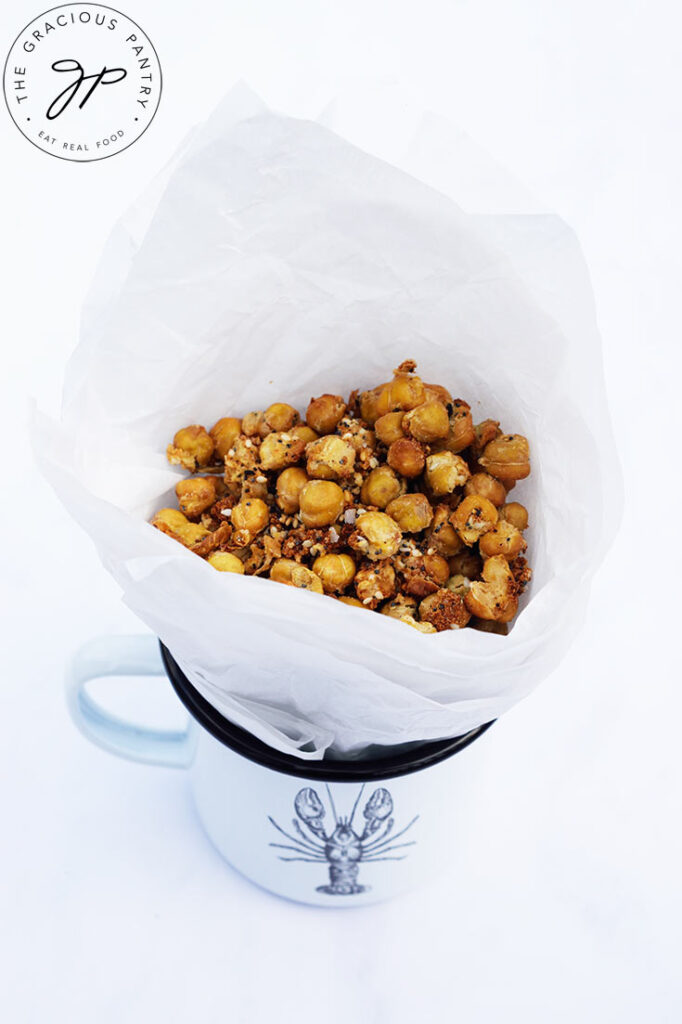 A white mug with a black lobster print sits on a white background holding a white parchment cone filled with Roasted Everything Bagel Chickpeas.