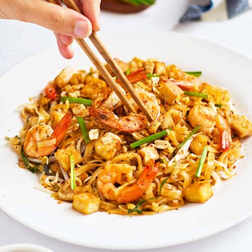 A female hand uses chopstick to pick up a shrimp from a plate of Shrimp Pad Thai.