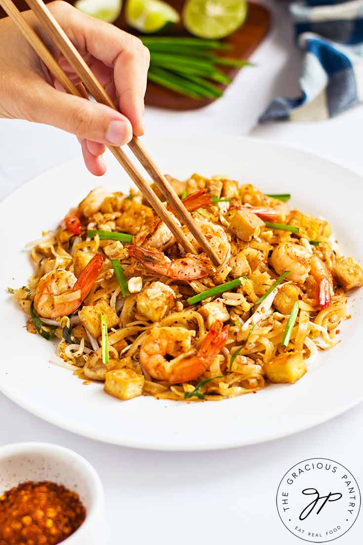 A female hand uses chopstick to pick up a shrimp from a plate of Shrimp Pad Thai.