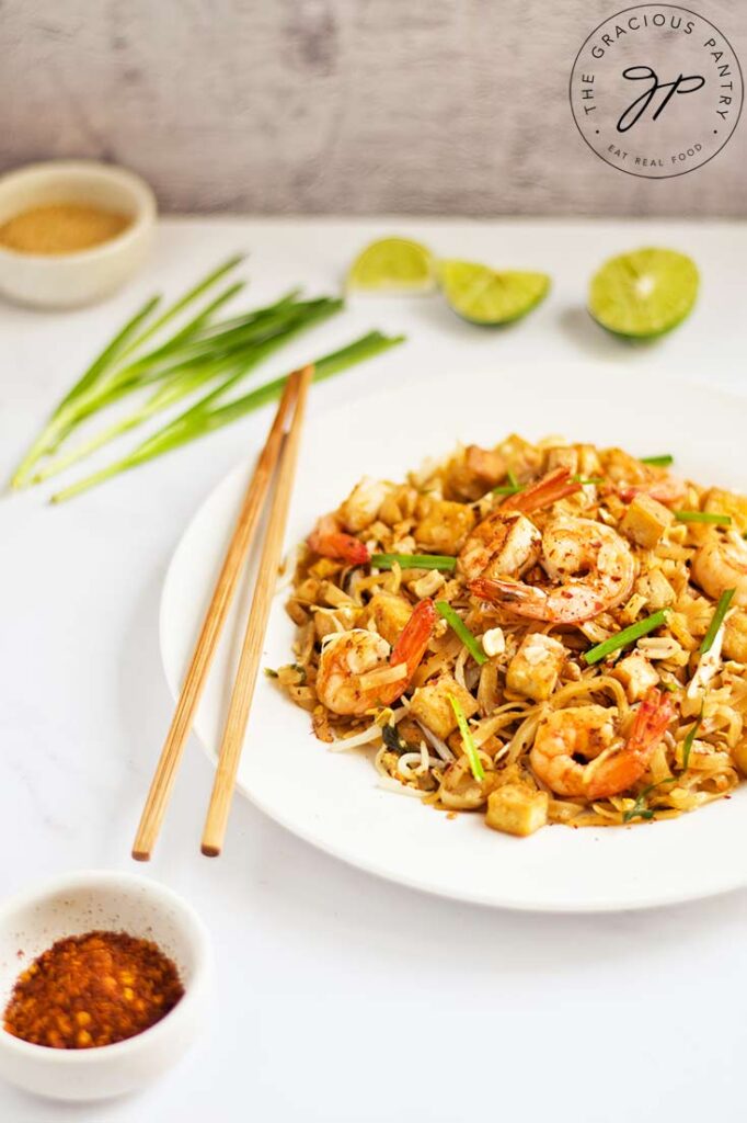 A partial view of a white plate holding Shrimp Pad Thai and chopseticks sits on a white surface.