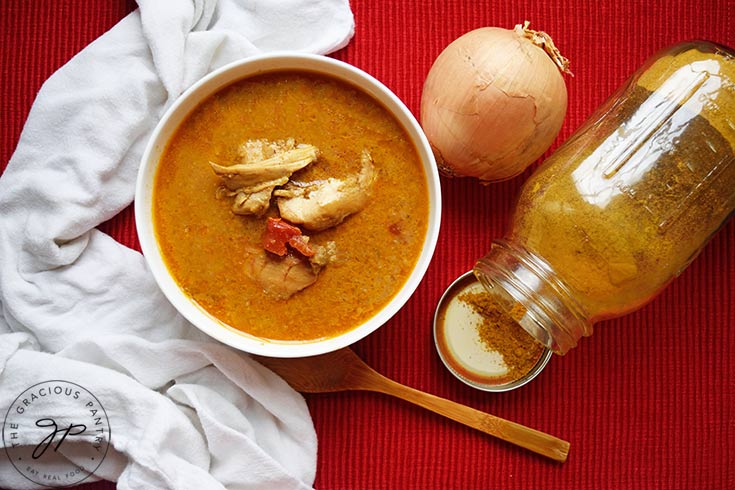 An overhead view of a white bowl filled with chicken curry soup. A jar of curry spice lays on it's side next to the bowl.