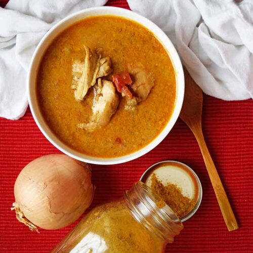 An overhead view of a white bowl of chicken curry soup, and yellow onion, a jar of curry powder and a wooden spoon laying on a red surface.