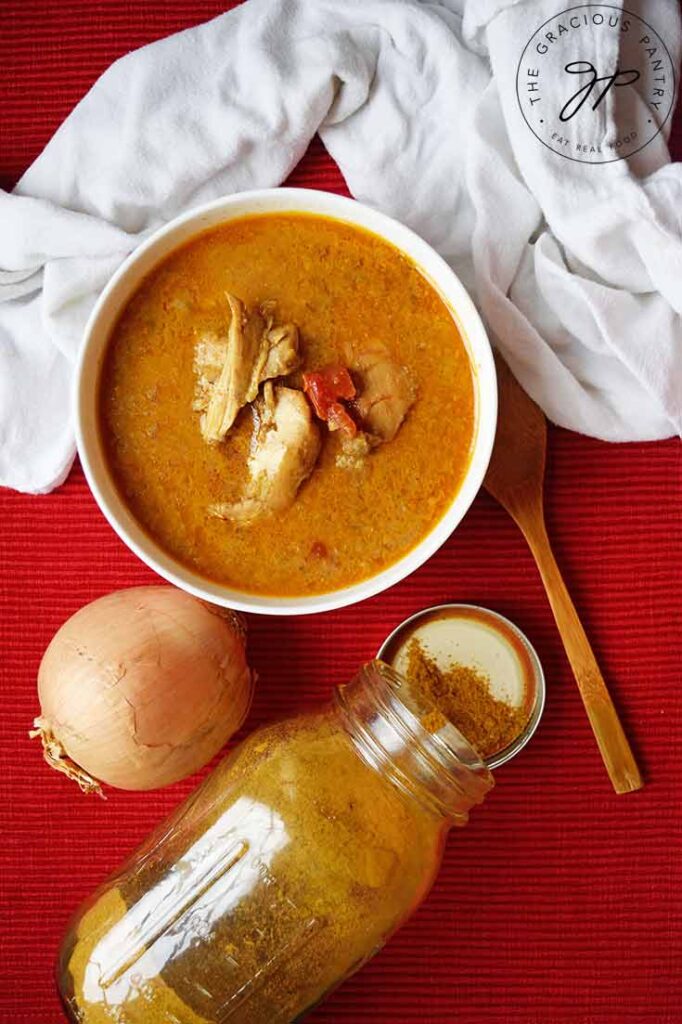 An overhead view of a white bowl of chicken curry soup, and yellow onion, a jar of curry powder and a wooden spoon laying on a red surface.