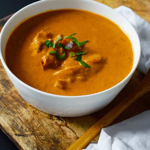A white bowl sits filled with butter chicken. A wooden spoon and cloth napkin lay to the side.